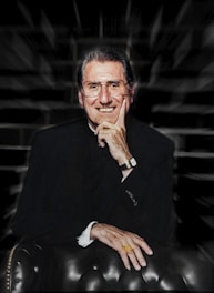 A smiling man in a dark suit, wearing glasses and a gold ring, is seated on a leather chair with his hand resting on his chin. The background is blurred with a zooming effect.