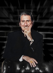 A smiling man in a dark suit, wearing glasses and a gold ring, is seated on a leather chair with his hand resting on his chin. The background is blurred with a zooming effect.
