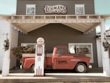 A vintage red pickup truck is parked against a building with a sign reading 'Lehman's.' An old-fashioned gas pump labeled 'Fleet-Wing' stands in front of the truck. The setting appears to be decorated for the holidays with greenery and red berries adorning the area.