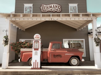 A vintage red pickup truck is parked against a building with a sign reading 'Lehman's.' An old-fashioned gas pump labeled 'Fleet-Wing' stands in front of the truck. The setting appears to be decorated for the holidays with greenery and red berries adorning the area.