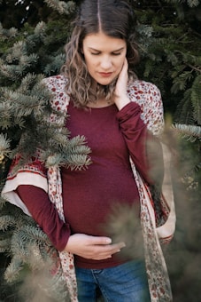 A woman stands amidst green pine branches with a serene expression, dressed in a maroon top and jeans, gently resting her hand on her belly, suggesting pregnancy. Her hair is wavy and she wears a patterned shawl.