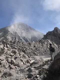 A rugged mountainous landscape with steep rocky slopes and a peak partially obscured by mist or clouds. A person with a backpack and outdoor gear stands in the foreground, gazing towards the mountain.
