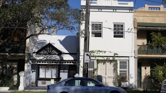 A row of terraced houses with distinct architectural styles is seen, featuring decorative facades and balconies. A car is parked on the street in front of the houses, and a road sign indicates a turn. Trees partially shade the scene, creating a dappled light effect.
