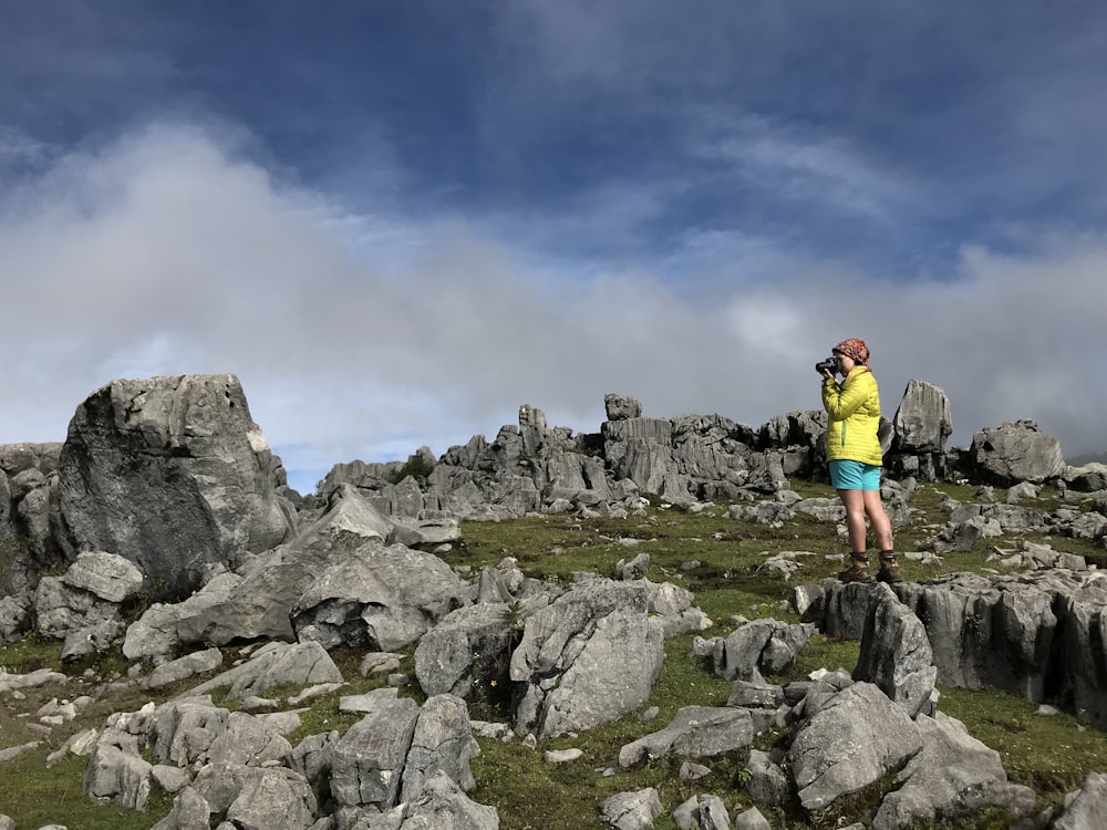 man in red t-shirt and black shorts standing on rocky mountain under white clouds during