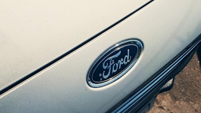 A close-up of a Ford car emblem displayed prominently on the vehicle's exterior. The oval-shaped emblem is surrounded by a chrome outline, set against the glossy surface of the car. The background has a smooth, metallic texture.