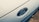 A close-up of a Ford car emblem displayed prominently on the vehicle's exterior. The oval-shaped emblem is surrounded by a chrome outline, set against the glossy surface of the car. The background has a smooth, metallic texture.