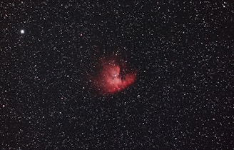 A vast expanse of the night sky with numerous small white stars scattered throughout the dark backdrop. At the center is a prominent, glowing red nebula with intricate details and varying intensity of light, surrounded by thin wisps of darker matter.