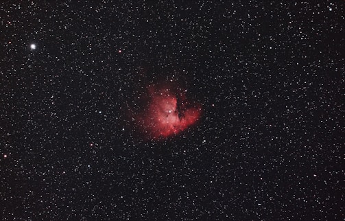 A vast expanse of the night sky with numerous small white stars scattered throughout the dark backdrop. At the center is a prominent, glowing red nebula with intricate details and varying intensity of light, surrounded by thin wisps of darker matter.