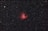 A vast expanse of the night sky with numerous small white stars scattered throughout the dark backdrop. At the center is a prominent, glowing red nebula with intricate details and varying intensity of light, surrounded by thin wisps of darker matter.