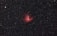 A vast expanse of the night sky with numerous small white stars scattered throughout the dark backdrop. At the center is a prominent, glowing red nebula with intricate details and varying intensity of light, surrounded by thin wisps of darker matter.