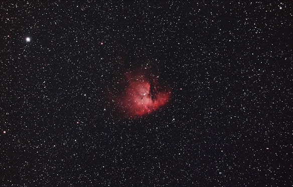 A vast expanse of the night sky with numerous small white stars scattered throughout the dark backdrop. At the center is a prominent, glowing red nebula with intricate details and varying intensity of light, surrounded by thin wisps of darker matter.