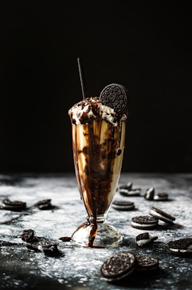A tall glass is filled with a creamy chocolate milkshake, topped with whipped cream, chocolate drizzle, and a single Oreo cookie. A black straw is inserted into the glass. The background is a dark, blurred setting with several Oreo cookies scattered around the glass on a textured surface.
