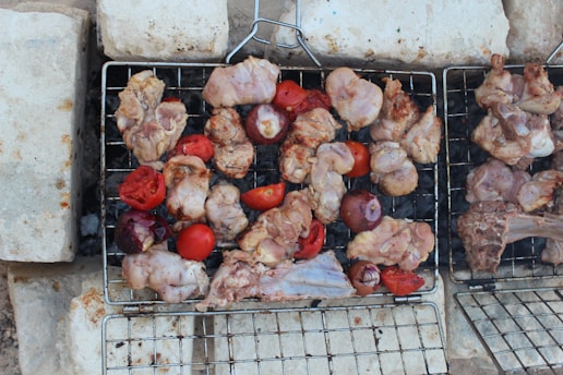 Pieces of raw chicken and tomatoes placed on a grill, surrounded by bricks, possibly preparing for a barbecue.