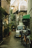 A narrow alleyway is flanked by various urban structures, including a small balcony and a green awning. A compact car is parked by the side, partially surrounded by potted plants and bicycles. The scene is lined with greenery, suggesting an urban garden setting.