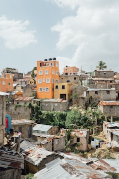A densely packed urban area with a mix of concrete and makeshift buildings, featuring a prominent orange multi-story building with water tanks on the roof. The area is surrounded by lush greenery and several homes with rusted metal roofs. Some laundry is visible drying in the sun.