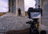 A digital camera is set up on a tripod, facing a person standing against a large concrete structure. The ground consists of cobblestone paving, and the sky is overcast. The person's figure is faintly visible on the camera's screen, indicating a photoshoot or filming session.