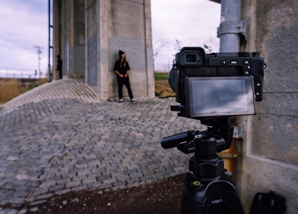 A digital camera is set up on a tripod, facing a person standing against a large concrete structure. The ground consists of cobblestone paving, and the sky is overcast. The person's figure is faintly visible on the camera's screen, indicating a photoshoot or filming session.
