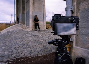 A digital camera is set up on a tripod, facing a person standing against a large concrete structure. The ground consists of cobblestone paving, and the sky is overcast. The person's figure is faintly visible on the camera's screen, indicating a photoshoot or filming session.