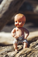 A vintage plastic baby doll is placed on rough wooden logs, its skin slightly faded with age. The doll wears a white cloth diaper fastened with a pin and has distinct facial features with blue eyes and orange lips.