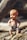 A vintage plastic baby doll is placed on rough wooden logs, its skin slightly faded with age. The doll wears a white cloth diaper fastened with a pin and has distinct facial features with blue eyes and orange lips.