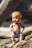 A vintage plastic baby doll is placed on rough wooden logs, its skin slightly faded with age. The doll wears a white cloth diaper fastened with a pin and has distinct facial features with blue eyes and orange lips.