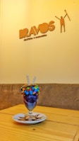 A colorful dessert glass filled with a layered dessert, topped with assorted candies and two wafer sticks, placed on a white plate on a wooden table. The background shows a wall with a logo that reads 'BRAVOS' accompanied by a stylized figure and additional text underneath.