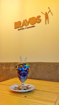 A colorful dessert glass filled with a layered dessert, topped with assorted candies and two wafer sticks, placed on a white plate on a wooden table. The background shows a wall with a logo that reads 'BRAVOS' accompanied by a stylized figure and additional text underneath.