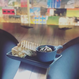 A grilled sandwich is placed next to a small cup filled with a garnish on a rectangular black plate. The setting is on a wooden table with a blurred colorful background, possibly an artistic wall or decor.