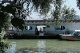 A calm river scene featuring a houseboat with a tiled roof and hexagonal windows. The houseboat is adorned with plants and set against lush green foliage. A small motorboat is docked beside the houseboat, with reflections visible on the water.