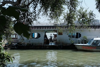 A calm river scene featuring a houseboat with a tiled roof and hexagonal windows. The houseboat is adorned with plants and set against lush green foliage. A small motorboat is docked beside the houseboat, with reflections visible on the water.