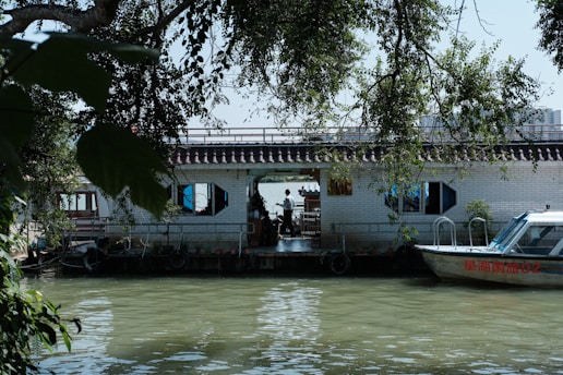 A calm river scene featuring a houseboat with a tiled roof and hexagonal windows. The houseboat is adorned with plants and set against lush green foliage. A small motorboat is docked beside the houseboat, with reflections visible on the water.