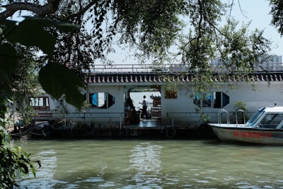 A calm river scene featuring a houseboat with a tiled roof and hexagonal windows. The houseboat is adorned with plants and set against lush green foliage. A small motorboat is docked beside the houseboat, with reflections visible on the water.