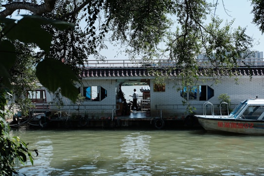 A calm river scene featuring a houseboat with a tiled roof and hexagonal windows. The houseboat is adorned with plants and set against lush green foliage. A small motorboat is docked beside the houseboat, with reflections visible on the water.