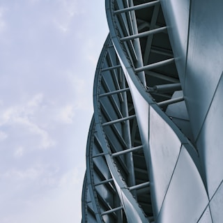 Curved, metallic architectural structure with overlapping panels and beams against a partly cloudy sky.