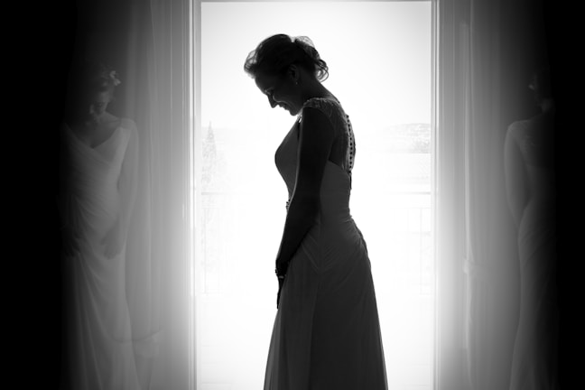A silhouette of a woman in a bridal gown is captured against a bright window backdrop, with curtains framing her graceful pose. The soft focus adds a dreamy quality to the image.
