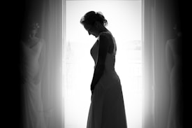 A silhouette of a woman in a bridal gown is captured against a bright window backdrop, with curtains framing her graceful pose. The soft focus adds a dreamy quality to the image.