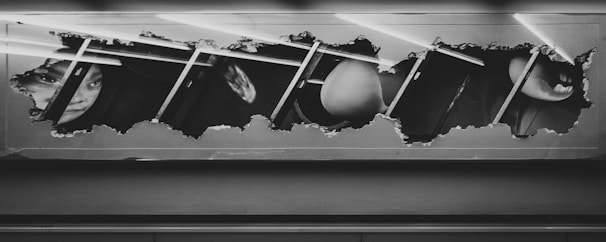 A rectangular frame with a ripped or torn effect revealing an underlying image of fragmented faces and objects, possibly glimpses of a person's face and other abstract shapes. The composition is monochromatic, predominantly in shades of black and white, with some diagonal lines crossing the image.