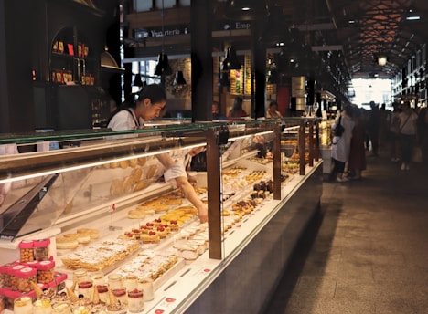 A person serves pastries and desserts displayed in a long glass counter at a bustling indoor market. Some of the sweets include tarts and cakes, and there are various jars and containers arranged neatly. The market is busy, with several people gathered nearby, illuminated by industrial-style overhead lighting.