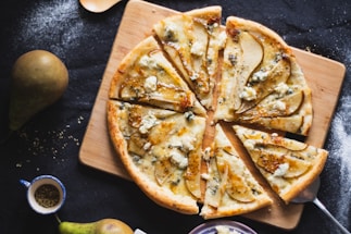 A gourmet pizza topped with thinly sliced pears and crumbled blue cheese is laid out on a wooden board. The pizza is cut into eight slices, showing a golden-brown crust and a creamy cheese base. Surrounding the pizza are whole pears, a small cup containing herbs, and scattered flour on a dark surface.