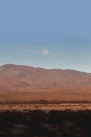 A serene landscape featuring a vast desert plain with scattered shrubs and bushes, leading up to a mountain range in the background. Above the mountains, a clear blue sky is accented with a faint, visible moon.