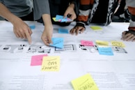 Several individuals are using colorful sticky notes on a table covered with printed papers. The notes have words like 'customer', 'problem', and 'journey map', indicating a collaborative planning or brainstorming session. Hands are visible, actively arranging the notes.