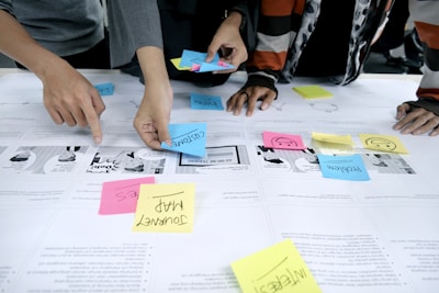 Several individuals are using colorful sticky notes on a table covered with printed papers. The notes have words like 'customer', 'problem', and 'journey map', indicating a collaborative planning or brainstorming session. Hands are visible, actively arranging the notes.