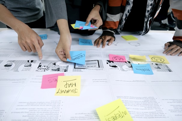 Several individuals are using colorful sticky notes on a table covered with printed papers. The notes have words like 'customer', 'problem', and 'journey map', indicating a collaborative planning or brainstorming session. Hands are visible, actively arranging the notes.