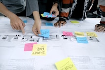 Several individuals are using colorful sticky notes on a table covered with printed papers. The notes have words like 'customer', 'problem', and 'journey map', indicating a collaborative planning or brainstorming session. Hands are visible, actively arranging the notes.