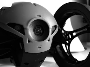 A close-up view of a robotic device, featuring a prominent lens or camera at its center. The design includes smooth, curved surfaces with a mix of futuristic elements. In the background, a circular, wheel-like structure adds depth to the composition.