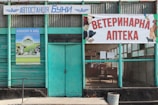 A building facade with a teal green color scheme, featuring a sign in Cyrillic script indicating a veterinary pharmacy. The entrance has a glass window and a wooden door. Posters with various animals, including a cow and dog, decorate the exterior.