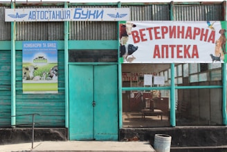 A building facade with a teal green color scheme, featuring a sign in Cyrillic script indicating a veterinary pharmacy. The entrance has a glass window and a wooden door. Posters with various animals, including a cow and dog, decorate the exterior.