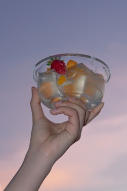 A hand is holding a glass bowl filled with clear jelly and pieces of fruit, including a strawberry and orange segments. The background is a soft, pastel-colored sky, suggesting a serene and calm atmosphere.