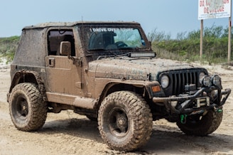 A muddy off-road vehicle, likely a Jeep, is parked on a dirt or sandy path. The vehicle is covered in dirt, indicating it has been used in rough terrain. It has large, rugged tires suitable for off-roading and additional equipment on the front, such as a winch and extra lights. The surrounding area is sparse with some greenery and a sign in the background.
