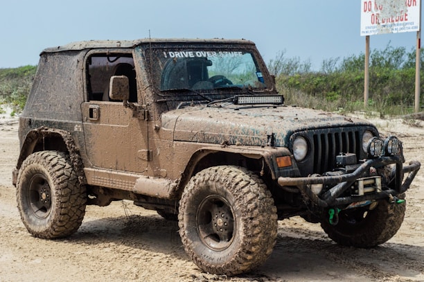 A muddy off-road vehicle, likely a Jeep, is parked on a dirt or sandy path. The vehicle is covered in dirt, indicating it has been used in rough terrain. It has large, rugged tires suitable for off-roading and additional equipment on the front, such as a winch and extra lights. The surrounding area is sparse with some greenery and a sign in the background.